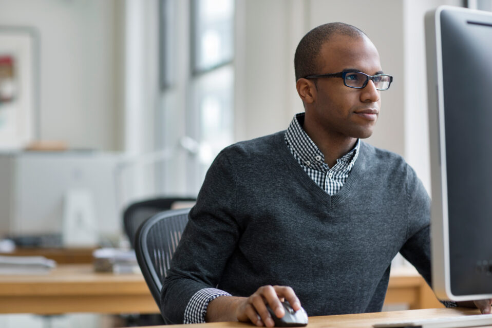 A man sits in front of a computer screen using a mouse. He has glasses and a gray sweater on.