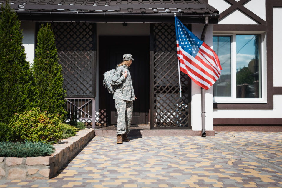 Military woman with backpack leaving house. An American flag is next to her.